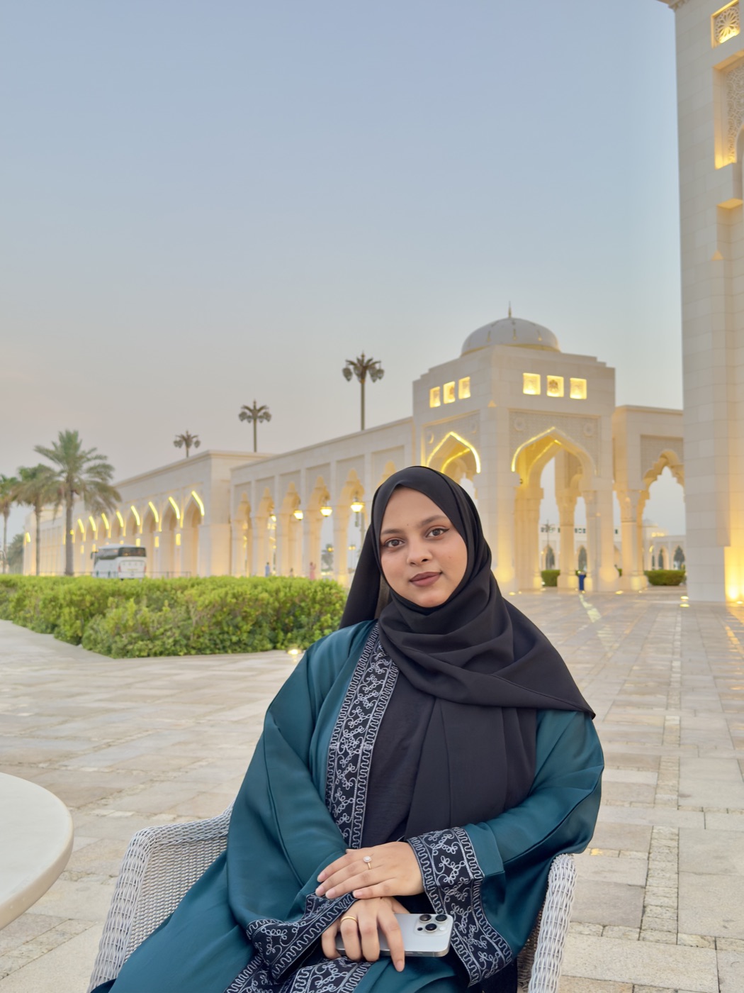 Jabin Akhter Borah seated before the white arcades of a mosque at dusk, in a teal abaya and black hijab