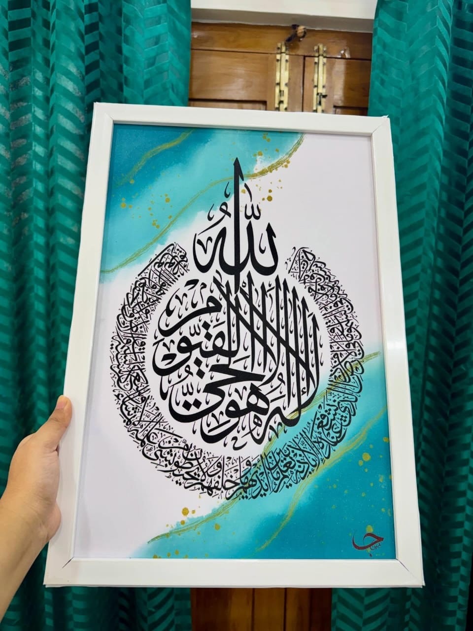 The artist's hand holding a white-framed Ayat al-Kursi painting over a turquoise wash