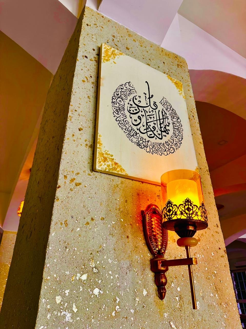A Jabin Arts calligraphy panel glowing beside a lantern on a mosque pillar at dusk