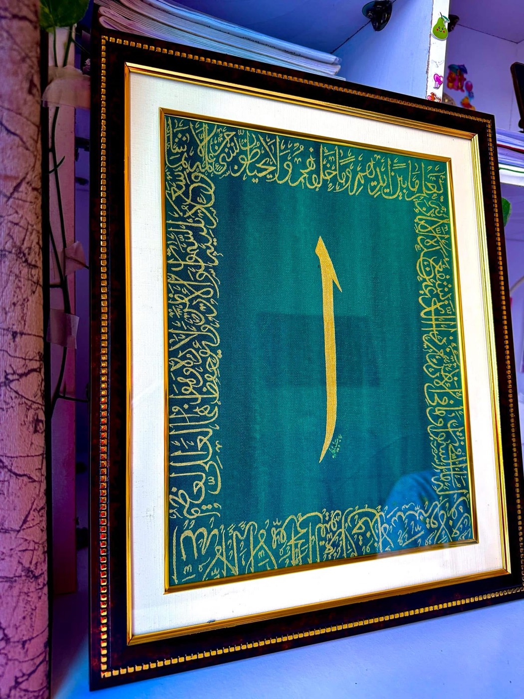 A single golden alif at the center of a teal panel bordered by gold script, ornate frame
