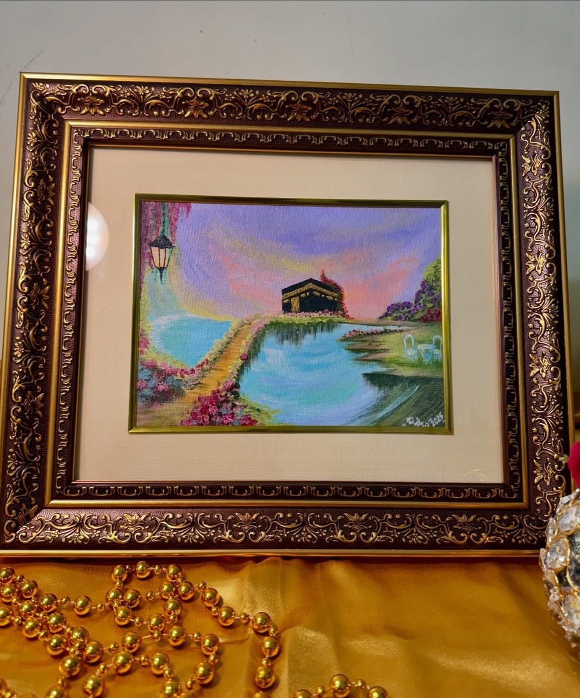 Framed painting of the Kaaba on a green islet above a turquoise lake, reached by a flowered path under a pastel sunset, in an ornate gilt frame