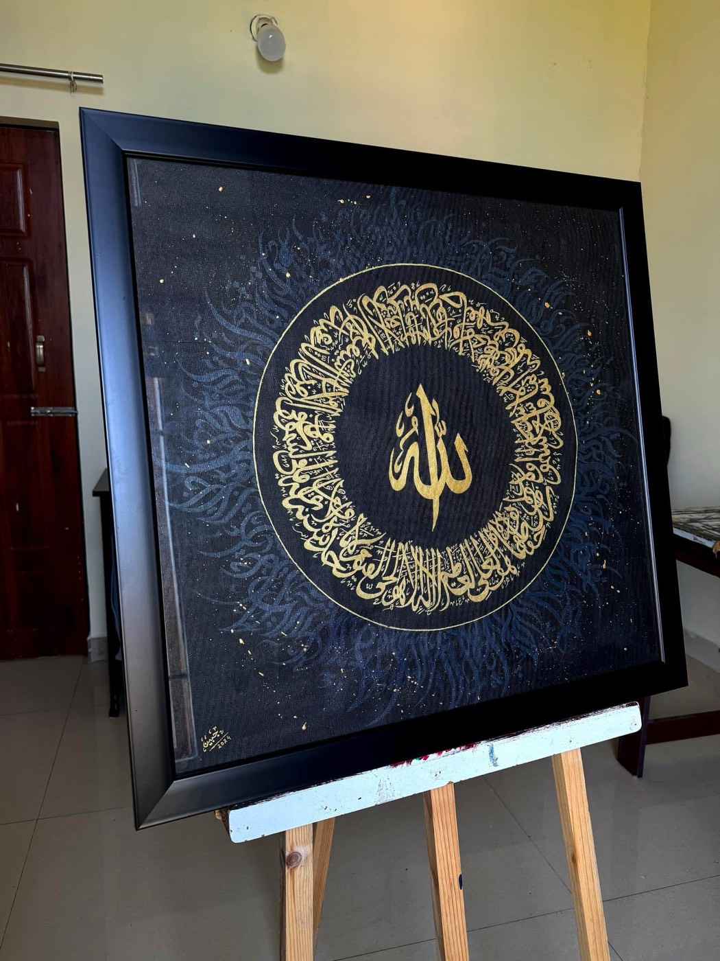 Dense circular gold script ring around the name Allah on black canvas, standing on an easel
