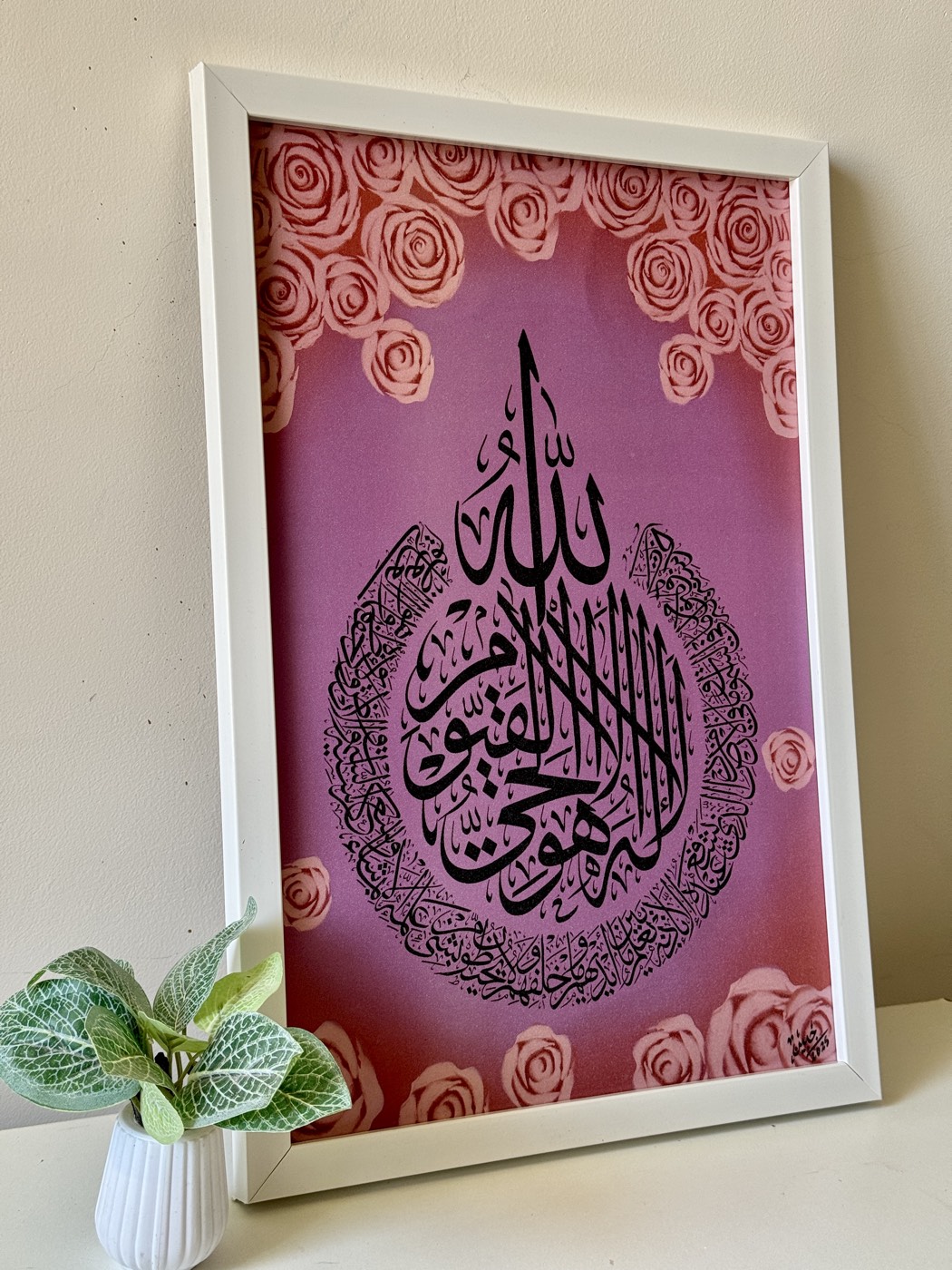 Ayat al-Kursi in black ink on a blush pink ground bordered with roses, white frame beside a plant