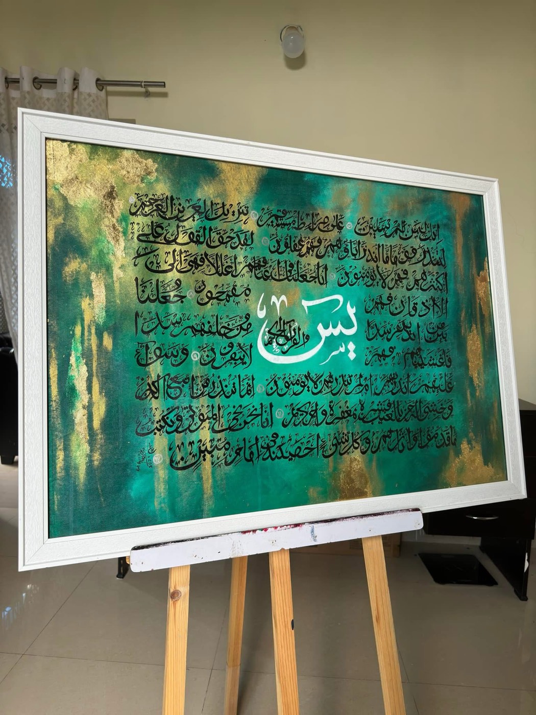 Wide painting of Surah Ya-Sin text over teal and gold mists, white frame on an easel
